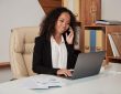 Professional woman in a suit talking on a smartphone while working on a laptop at a cluttered office desk with papers nearby