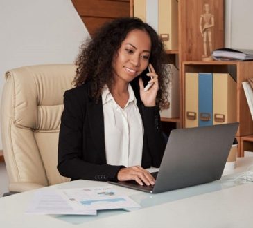 Professional woman in a suit talking on a smartphone while working on a laptop at a cluttered office desk with papers nearby