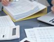 Person using a calculator on a desk filled with financial papers and a laptop nearby.