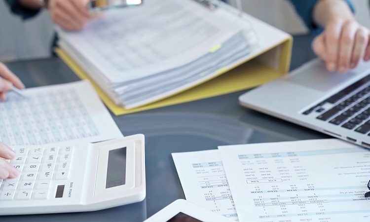 Person using a calculator on a desk filled with financial papers and a laptop nearby.