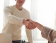 Two people shake hands over a desk in a bright room, signaling a greeting or agreement.