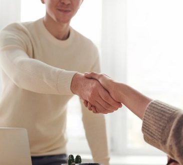 Two people shake hands over a desk in a bright room, signaling a greeting or agreement.