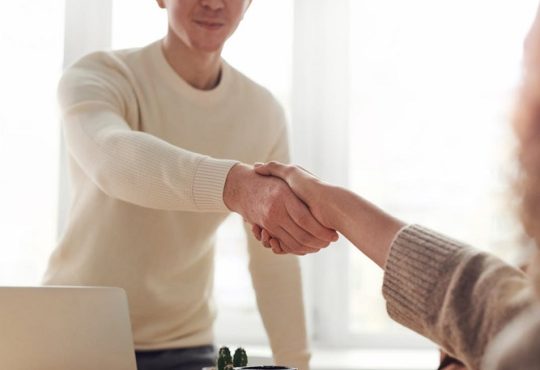 Two people shake hands over a desk in a bright room, signaling a greeting or agreement.