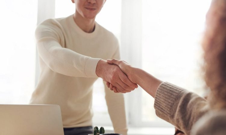 Two people shake hands over a desk in a bright room, signaling a greeting or agreement.