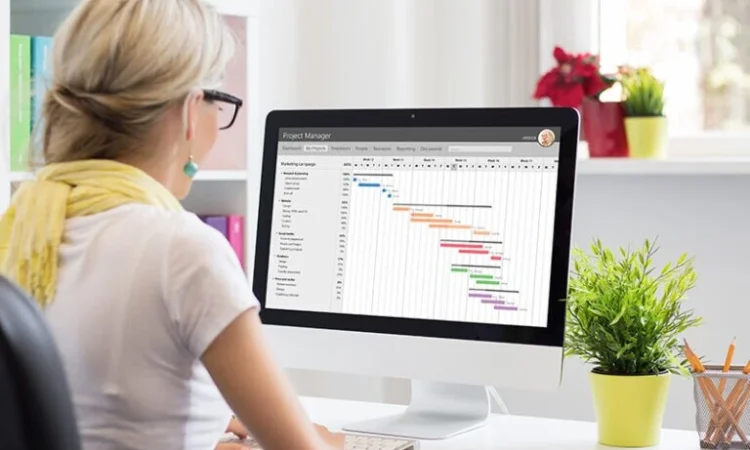 Woman at a bright desk reviewing a project management Gantt chart on a desktop monitor